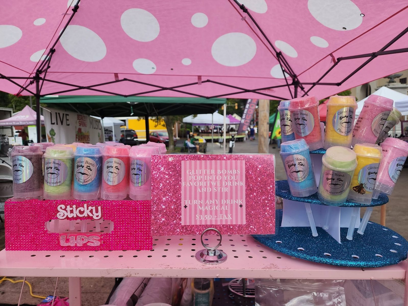 The Sticky Lips booth under a pink polka-dot canopy, with a rhinestone Sticky Lips display and rows of Glitter Bomb cotton candy tubes in grape, watermelon, blue raspberry, cherry and strawberry