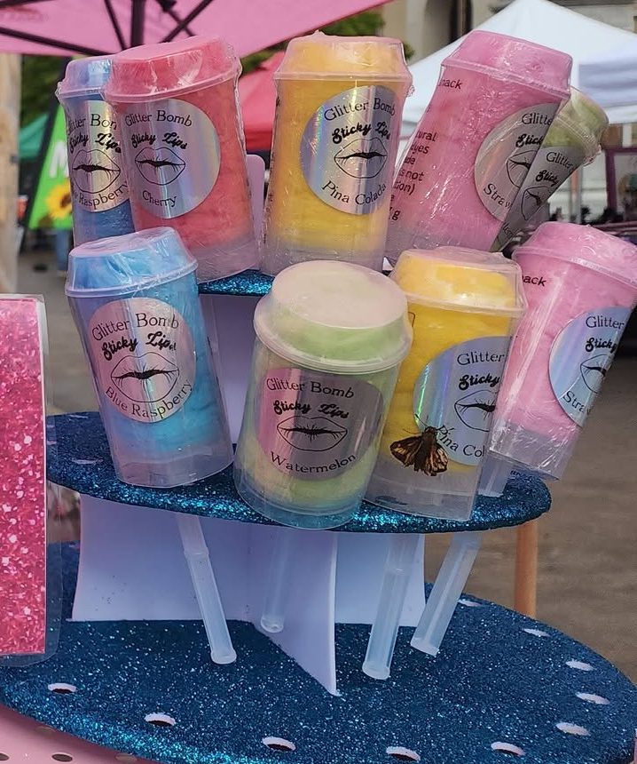 Glitter Bomb tubes stacked on a blue glitter stand, each labeled Sticky Lips with flavors including cherry, blue raspberry, pina colada, watermelon and strawberry